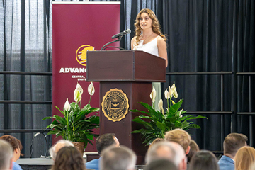 A woman in a white dress is on a platform behind a podium speaking to a seating crowd. White flowers are on both sides of the podium and a CMU Advancement banner, CMU flag and USA flag stands behind her on the stage.