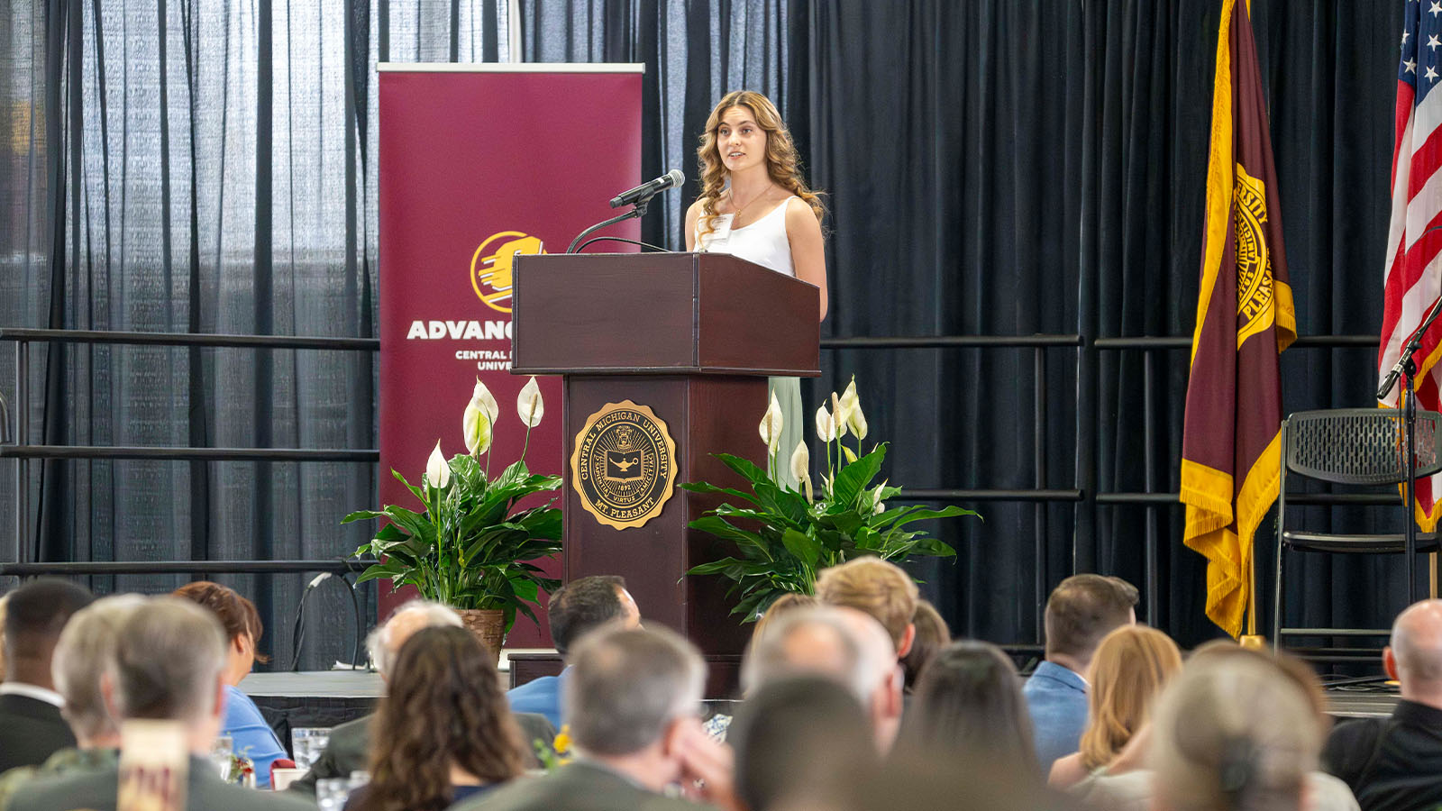 A woman in a white dress is on a platform behind a podium speaking to a seating crowd. White flowers are on both sides of the podium and a CMU Advancement banner, CMU flag and USA flag stands behind her on the stage.