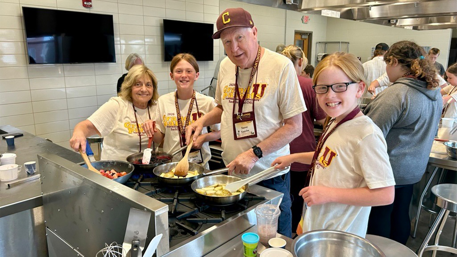 A group of four people, a senior citizen woman, a senior citizen man, and two younger girls, are cooking in a kitchen.