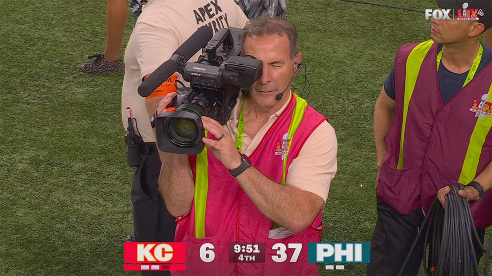 A man with a video camera is wearing a red vest with the NFL logo on the back during Fox's Super Bowl LIX broadcast.
