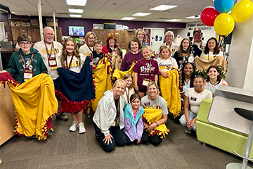 A group of people pose for a photo inside a large room with four balloons in the upper right corner. Some people are holding quilts in various colors like yellow, blue and tan.
