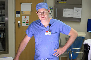 A man with glasses on is wearing scrubs inside a hospital room.