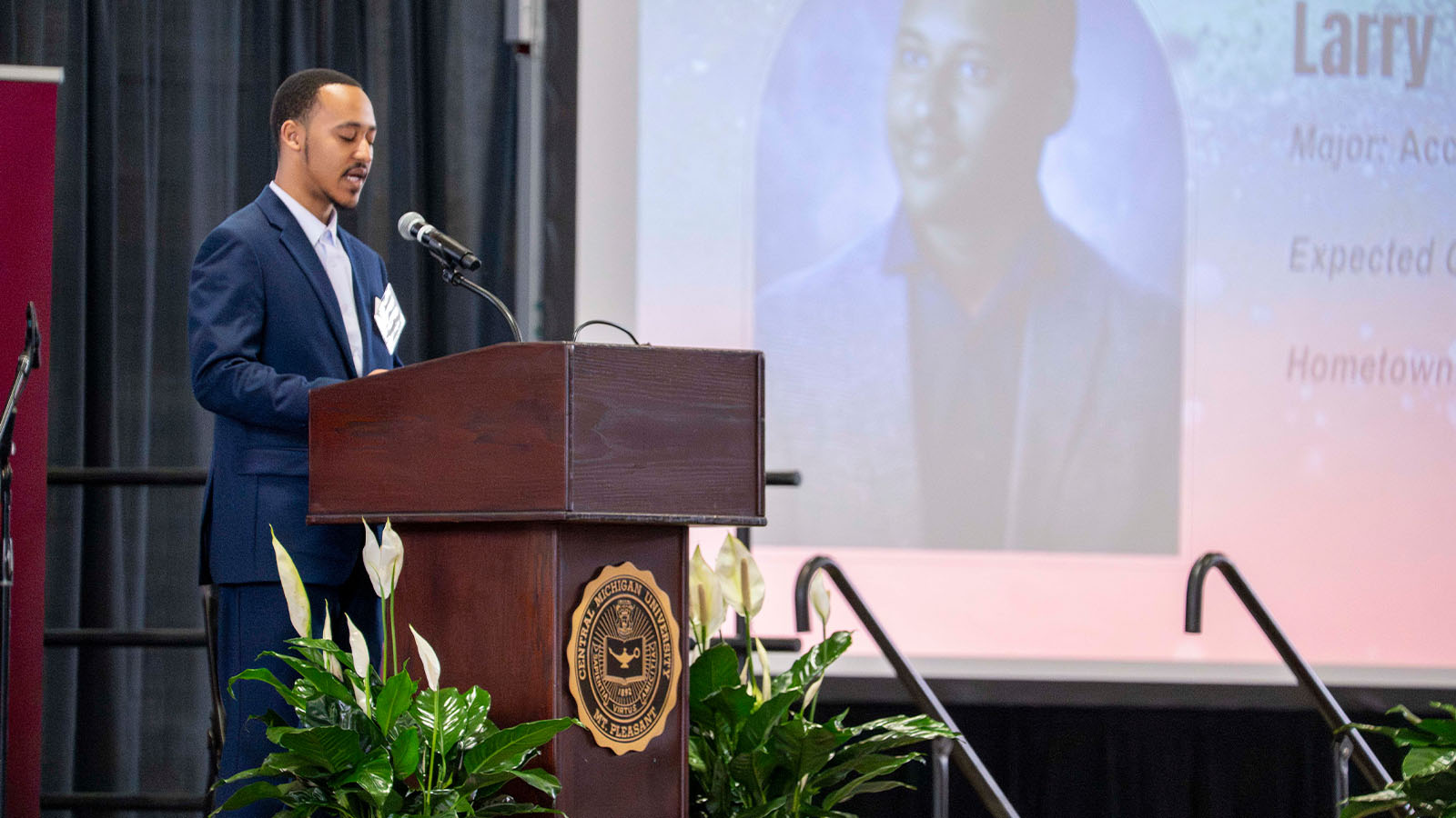 A man in a suit is speaking behind a podium with white flowers on both sides of the podium and a projected picture of himself in the background.