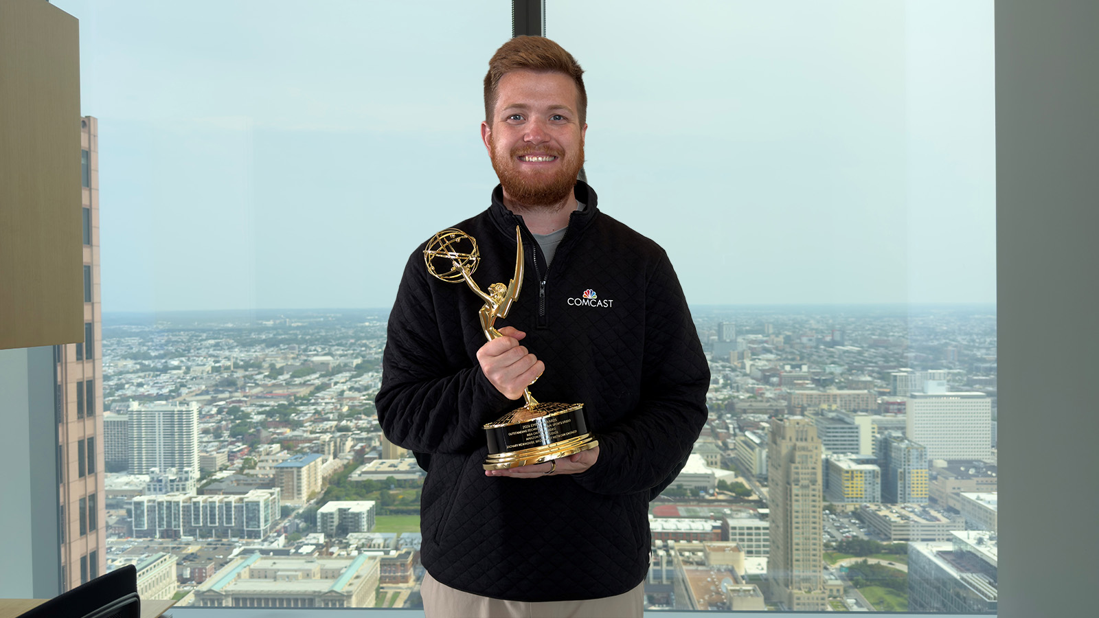 A man with a beard wearing a black quarter zip holds a trophy in front of a window with the view of a city in the background.