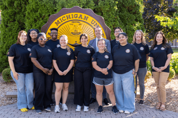a25-09-29 Compass mbh-3 - 600x400 Compass students and staff in black t-shirts posing with President MacKinnon in front of the CMU Seal.