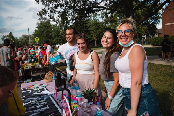 Students in white tank tops standing at a table outside with flyers and trinkets.