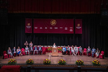 a group of people sitting on a stage adorned with flowers, maroon chairs, and maroon and gold banners